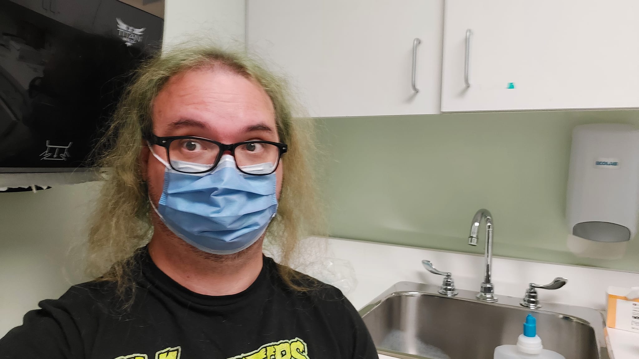 A man with long green hair sits in an old examnation room in a hospital